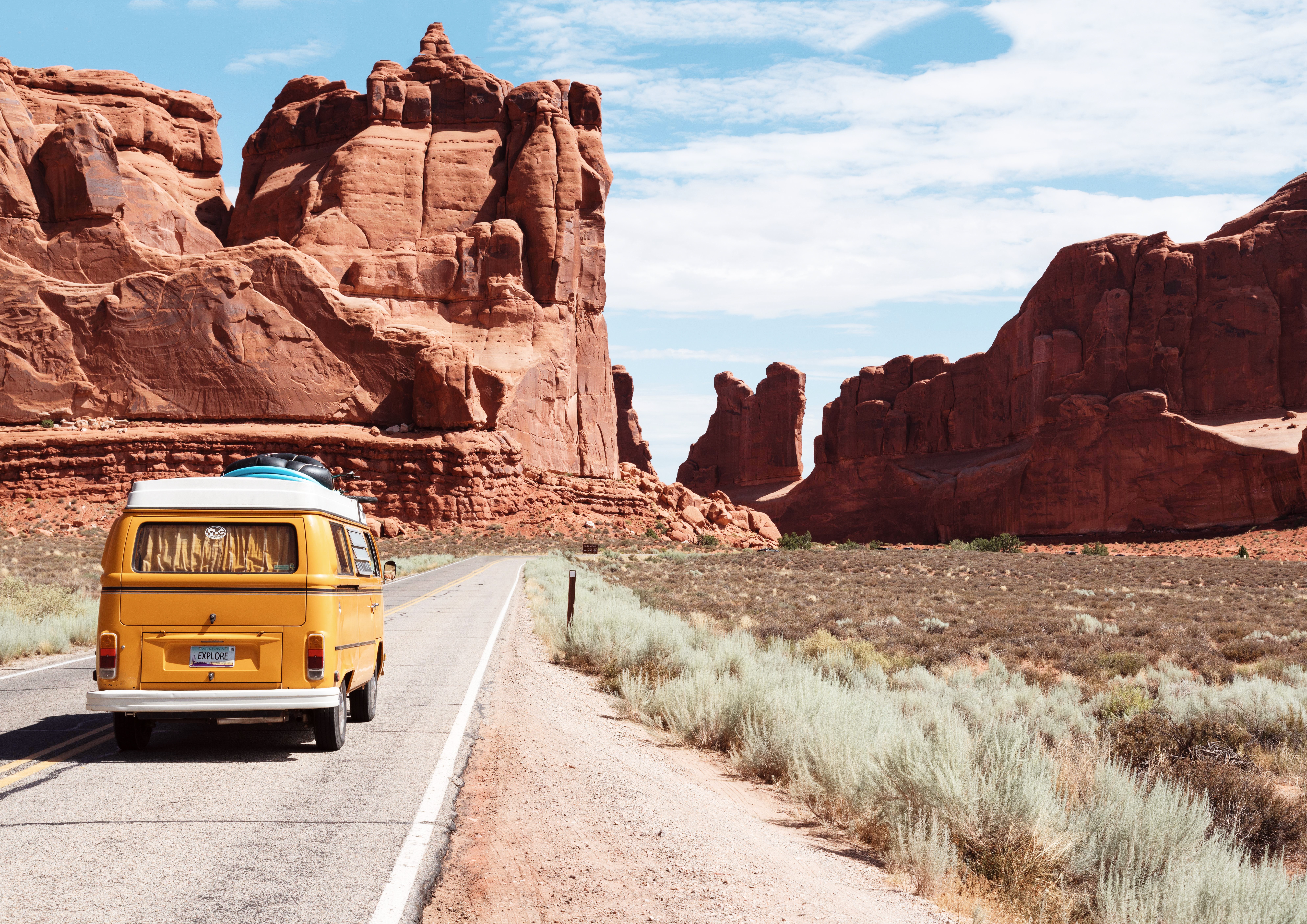 An old-fashioned 1970's camper van drives down an empty road surrounded by desert and sandy-coloured rocky mountains.